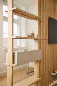 WoodUpp roomdivider van licht eikenhout: slanke open stelling met planken en verticale lamellen, zichtbaar vastgezet met RVS-bouten. Op een plank staat een grijze metalen WoodUpp kabelbak (kabelbox) en wat decoratief touw, naast een wand met houten latten en een tv.