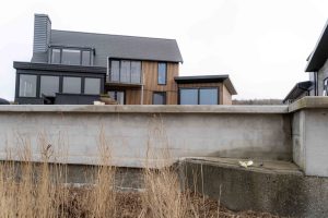 Voorgevel van modern huis met WoodUpp-gevel: smalle, verticale houten lamellen in lichtbruin die centrale gevel en aanbouw bekleden, gecombineerd met grote zwarte kozijnen en glazen veranda. Betonmuur en droge rietstengels op voorgrond; WoodUpp gevelbekleding en lamellenpanelen benadrukken de strakke architectuur.