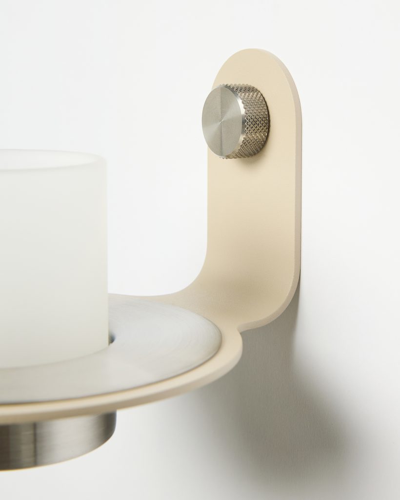 Close-up van een WoodUpp wandkandelaar: een beige metalen wandbeugel met afgeronde vorm en satijnfinish, een matwit glazen cilinder voor kaars of lamp, plus een geborstelde metalen ring en geruwde knop als bevestiging. Minimalistisch, functioneel Scandinavisch ontwerp met subtiele texturen en strakke lijnen.