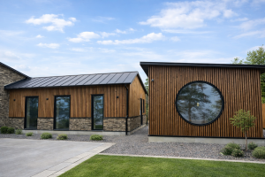 Een buitenaanzicht van moderne gebouwen waarvan de gevels grotendeels zijn bekleed met WoodUpp verticale houten latten in een donkere tint, waarschijnlijk gerookt eiken. De Akupanels worden gecombineerd met een natuurstenen plint en zwarte kozijnen, inclusief een opvallend groot rond raam in het rechtergebouw.