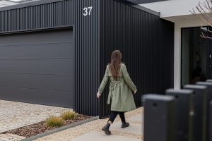 Vrouw loopt langs een moderne woning met zwarte verticale WoodUpp-gevellatten en een brede antraciet garagedeur; op de voorgrond wazig een zwart WoodUpp-lattenhek. Strakke, matte lamellen en scherpe hoeken benadrukken het minimalistische exterieur, huisnummer 37 zichtbaar, vooraan grindpad met lage beplanting.