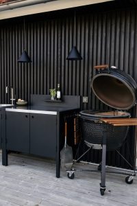 Outdoor keuken tegen een zwart houten gevelwand met een WoodUpp matzwarte werkbank/keukenkast met spoelbak, roestvrijstalen rand, opberglades en klein opbergplankje; twee zwarte hanglampen erboven en een open kamado-grill met houten handvat en opklapbare zijplank op een houten vlonder, met een fles olie en een klein vaasje basilicum.